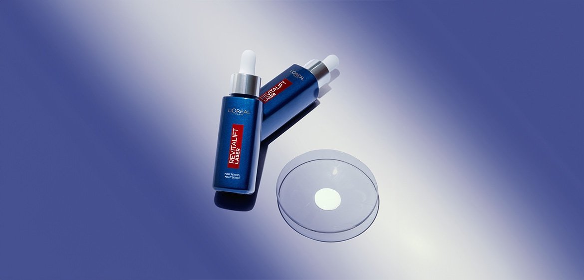 Zwei Fläschchen Revitalift Laser Pure Retinol Night Serum neben einer Glasschale mit einem Tropfen Serum vor einem violetten Hintergrund